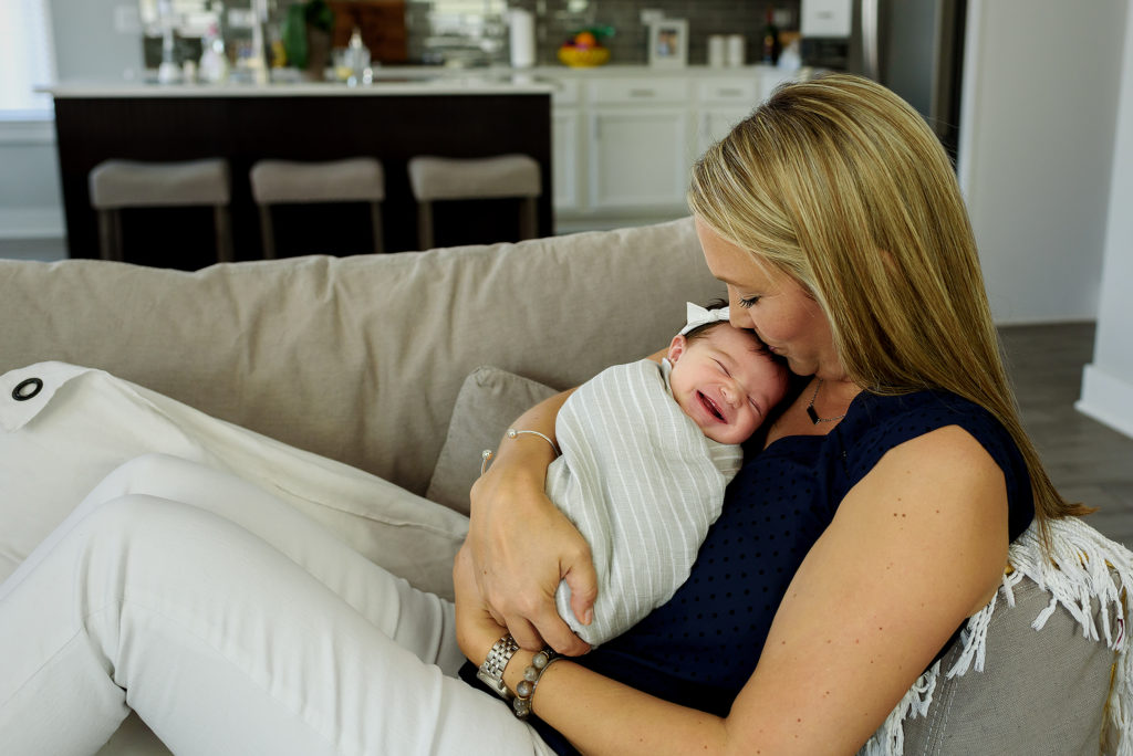 Mom holding and kissing smiling baby girl on couch for lifestyle newborn session in Jacksonville Beach, FL.