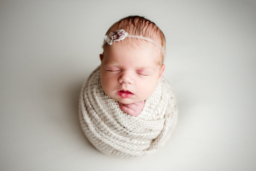 Newborn girl on neutral background in potato pose with floral headband in Jacksonville, FL.