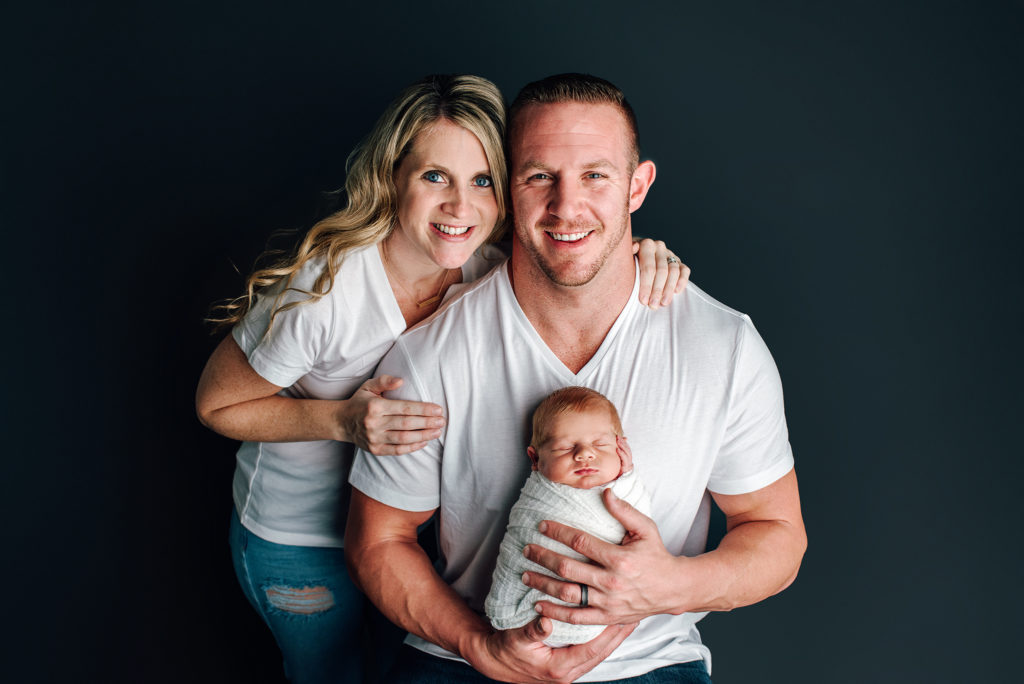 Parents wearing white shirt and jeans holding newborn boy for studio family session in Jacksonville Beach, FL.